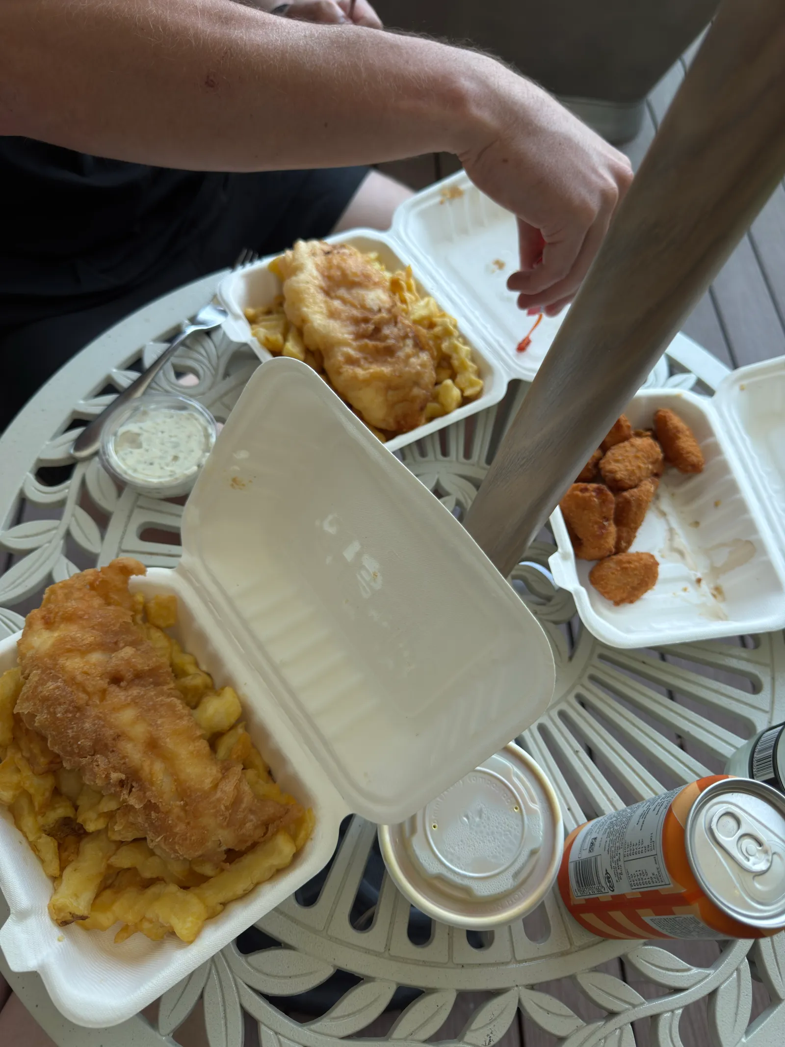 A photo taken from above of two boxes of fish and chips and a some fried scampi on a metal round outdoor table.